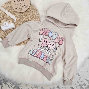 Lottie & Lysh product image featuring the stone sweat hooded sweatshirt with choose kindness printed graphic, against a white fluffy rug. Accenting the image are a wooden, toy camera and Lottie & Lysh wooden name plaque.