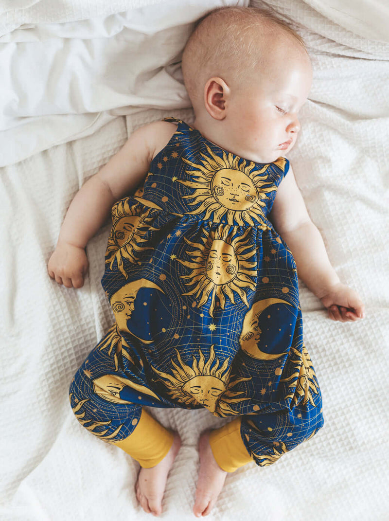 star and moon baby romper. Dark blue background with golden star and moon printed design. Worn by a baby girl lying on a white blanket.