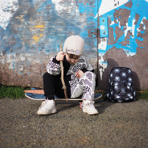 Toddler boy sitting on a skateboard against a graffiti wall. He is wearing a black and white animal print tracksuit with beige smile face beanie hat. On the floor next to him there is a black lion print toddler sized backpack.