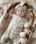 sleeping baby girl wearing a beige, floral romper in neutral tones with matching headband. she is sleeping in a bassinet with various knitted toys next to her.