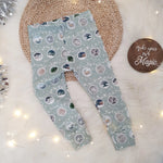Christmas bauble printed leggings by Lottie & lysh. These baby and child leggings feature a light green background with garlands of baubles each depicting a different wintery scene. The leggings are pictured against a white, fluffy rug with wicker placemat, silver tinsel and fairy lights to accent.