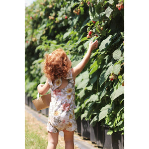 toddler girl with red curly hair reaching up to pick a raspberry from the bush. Wearing a bow back playsuit by Lottie & Lysh