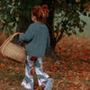 Red haired toddler girl wearing a green knitted jumper and flared trousers. She is carrying a wicker basket and the scene is autumnal with leaves on the ground and golden leaves on the trees.