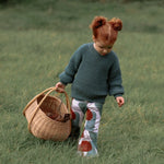Toddle girl walking through a field carrying a wicker basket. She has red hair in bunches, and is earing a green knitted jumper with printed flared trousers. 