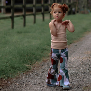 Young toddler girl with auburn hair walking down a gravel path. She is wearing printed toddler flared trousers and a peach coloured vest