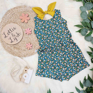 Product image for Lottie & Lysh. Featuring a green floral fabric with mustard accents in a girls summer romper with bow back detail. Pictured against a white fluffy backdrop with leaves to accentuate colours, branding disk and toy camera as props