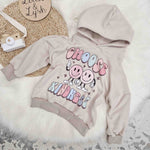 Lottie & Lysh product image featuring the stone sweat hooded sweatshirt with choose kindness printed graphic, against a white fluffy rug. Accenting the image are a wooden, toy camera and Lottie & Lysh wooden name plaque.