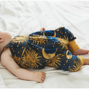 Young baby girl laying on a white bed, wearing a blue and gold boho romper accented with a 90s style sun and moon print