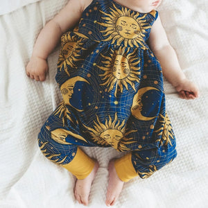 A baby girl wearing a 90s inspired, celestial print baby romper by Lottie & Lysh. The fabric is dark blue and features gold stars, moons and sunshines in a boho style.