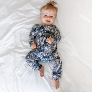 smiling baby boy pictured on a white bed, wearing an owl print babygro by Lottie & lysh