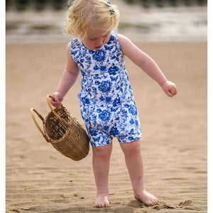A little girl with blonde hair playing on a beach. She is wearing a blue and white floral playsuit and is carrying a wicker basket in her hand
