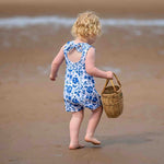 blonde toddler girl on a beach. She is wearing a bow back blue and white floral romper by Lottie & Lysh and carrying a wicker basket. She is walking away from the camera
